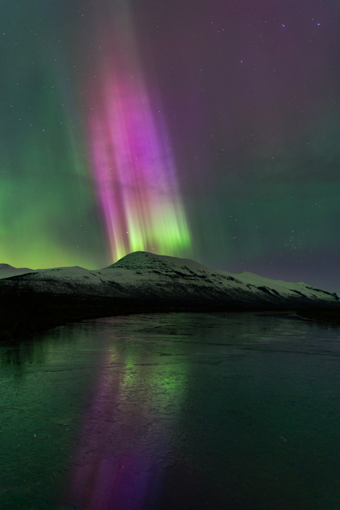 Aurora boreal de color lila sortint d'una muntanya i reflectida en un llac gelat a Nikkaluokta, Lapònia sueca.